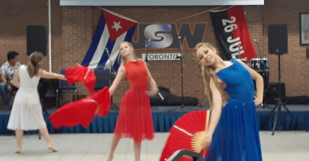 Three dancers in red, white and blue perform with fans beneath a Cuban flag and a 26 de Julio banner at CCFA Toronto's 2023 Moncada Day celebration at Steelworkers Hall